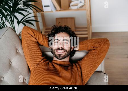 Glücklicher junger Mann, der sich zu Hause mit den Händen hinter dem Kopf auf dem Sofa entspannt Stockfoto