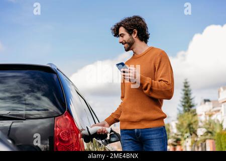 Lächelnder junger Mann, der das Auto an der Fahrzeugladestation auflädt Stockfoto