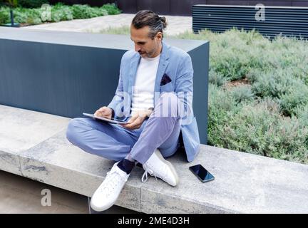 Ein erwachsener Mann, der einen Tablet-PC verwendet und auf einer Bank sitzt Stockfoto