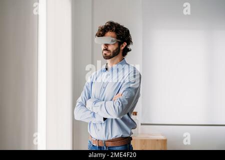 Ingenieur trägt eine VR-Brille und steht im Büro mit gekreuzten Armen Stockfoto