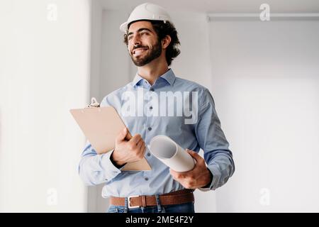 Glücklicher Ingenieur mit Schutzhelm und Bauplänen im Büro Stockfoto