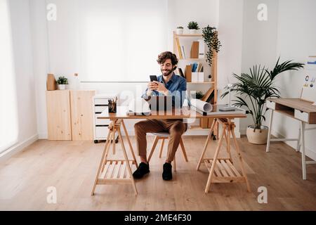 Glücklicher junger Ingenieur, der ein Mobiltelefon mit einem Laptop im Büro verwendet Stockfoto