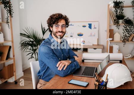 Glücklicher junger Ingenieur mit Brille, der mit einem Laptop am Schreibtisch im Büro sitzt Stockfoto