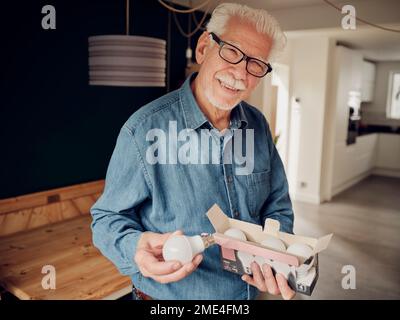 Ein älterer Mann, der zu Hause eine Schachtel LED-Glühbirnen in der Hand hat Stockfoto