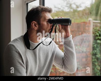 Ein Mann, der zu Hause mit einem Fernglas aus dem Fenster schaut Stockfoto