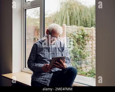 Seniorenmann am Fenster zu Hause mit einem digitalen Tablet Stockfoto