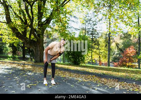 Der Läufer hat sich beim Joggen im Park in der Nähe der Bäume das Bein verletzt, der lateinamerikanische Mann hat Muskelkrämpfe, die sein Knie massieren, der Mann läuft und trainiert an einem sonnigen Tag. Stockfoto
