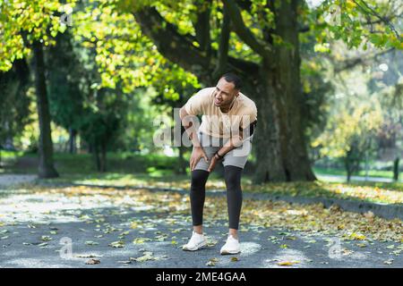 Der Läufer hat sich beim Joggen im Park in der Nähe der Bäume das Bein verletzt, der lateinamerikanische Mann hat Muskelkrämpfe, die sein Knie massieren, der Mann läuft und trainiert an einem sonnigen Tag. Stockfoto