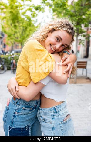 Lächelnde junge Frau mit blondem Haar, die eine Freundin auf dem Fußweg umarmt Stockfoto