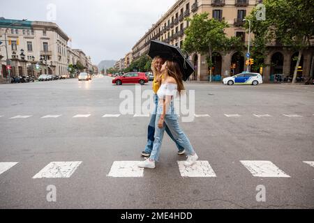 Glückliche junge Frau mit Schirm und Freundin, die die Straße in der Stadt überquert Stockfoto