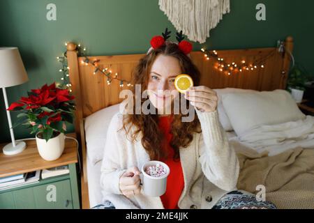 Lächelnde Frau mit einer Tasse heißer Schokolade und Marshmallows, die zu Hause auf dem Bett sitzt Stockfoto