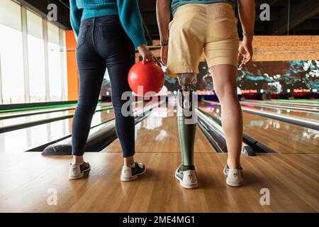 Eine Frau, die mit einer Freundin Ball hält und künstliche Gliedmaßen trägt, steht auf dem Parkettboden in der Bowlingbahn Stockfoto