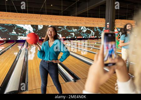 Eine Frau fotografiert eine Freundin mit einem Smartphone in einer Bowlingbahn Stockfoto