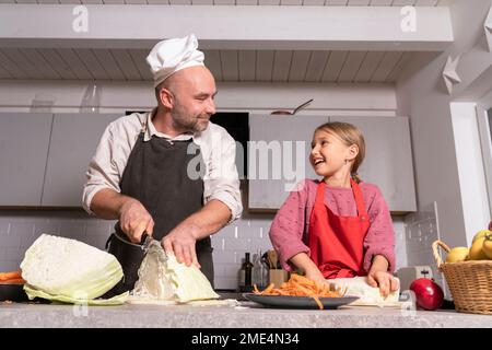 Vater und Tochter tragen Schürzen, die zu Hause in der Küche eine gesunde Mahlzeit zubereiten Stockfoto