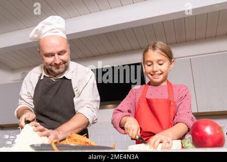 Vater und Tochter tragen Schürzen, die zu Hause in der Küche eine gesunde Mahlzeit zubereiten Stockfoto