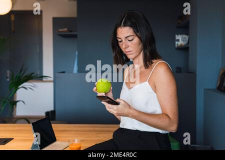 Junge Frau, die ein Smartphone benutzt und zu Hause Apfel hält Stockfoto
