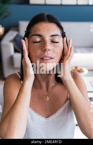 Frau mit geschlossenen Augen, die zu Hause Feuchtigkeitscreme auf das Gesicht aufträgt Stockfoto