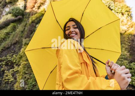 Lächelnde junge Frau mit gelbem Regenschirm vor dem vertikalen Garten Stockfoto