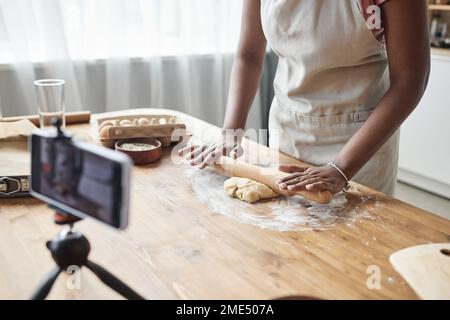 Nahaufnahme einer schwarzen Frau, die hausgemachtes Gebäck und rollenden Teig auf einer hölzernen Küchentheke backt, mit Smartphone-Kamera, die Video aufnimmt oder Livestream, Kopierbereich Stockfoto