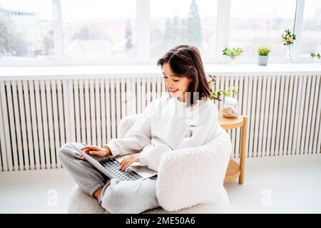 Lächelnde junge Frau, die zu Hause am Fenster sitzt und einen Laptop benutzt Stockfoto