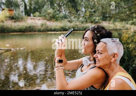 Junge Frau mit einer Freundin, die Selfie über Handy am Seeufer spricht Stockfoto