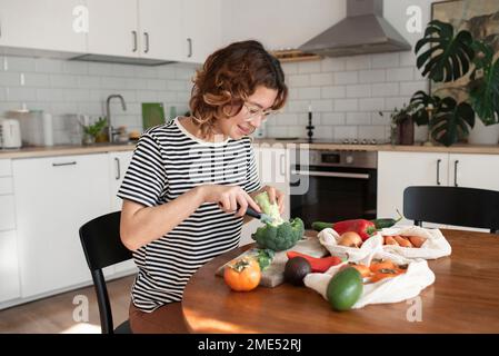 Lächelnde junge Frau, die Brokkoli schneidet, sitzt am Tisch in der Küche Stockfoto