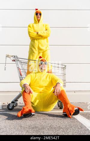 Eine Frau im Hühnerkostüm, die im Einkaufswagen steht, mit einem Mann, der auf dem Fußweg sitzt Stockfoto