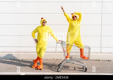 Mann und sorglose Frau in Hühnerkostümen stehen mit einem Einkaufswagen vor der Wand Stockfoto