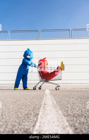 Ein Mann mit Kostüm schiebt eine Frau, die an sonnigen Tagen im Einkaufswagen sitzt Stockfoto