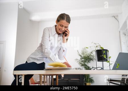 Glückliche, reife Geschäftsfrau, die im Büro Notizen schreibt und über ein Mobiltelefon spricht Stockfoto