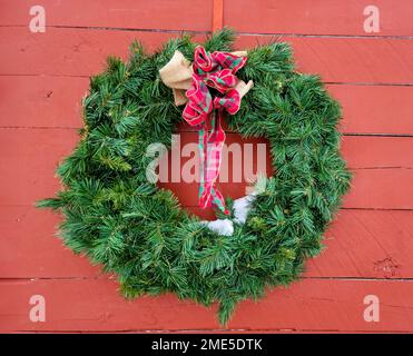 Grüner Weihnachtskranz mit einer karierten Schleife und schmelzendem Schnee auf einer roten Scheune Stockfoto