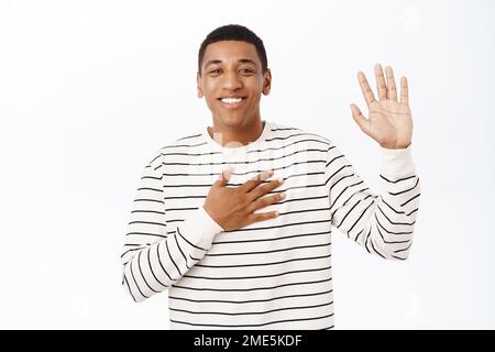 Lächelnder afroamerikanischer Mann stellt sich vor, hebt eine Hand und legt den Arm auf das Herz, begrüßt Sie, steht über weißem Hintergrund Stockfoto