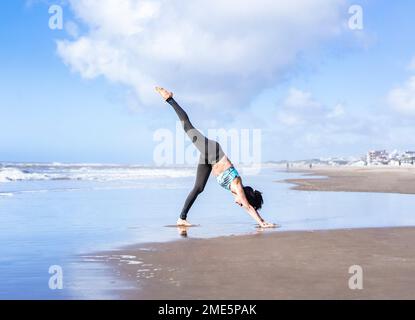 Eine Frau, die am Strand Yoga auf dem Wasser praktiziert und mit drei Beinen nach unten schauenden Hund (Eka Pada Adho Mukha Shvanasana) posiert. Stockfoto