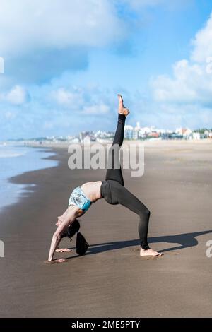 Eine Frau, die am Strand Yoga auf dem Wasser praktiziert und in einer einbeinigen Rad-Pose (Eka Pada Urdhva Dhanurasana) spielt. Stockfoto