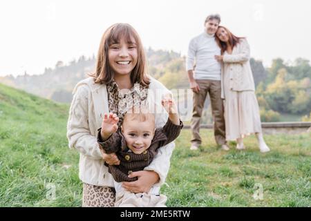 Eine weiße Teenagerin mit braunen Augen, dunklen Haaren und Pony in einem Kleid mit Leopardenmuster und einem Strickpullover hält ihren kleinen Bruder Kleinkind angezogen Stockfoto