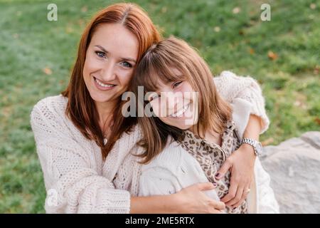 Eine junge weiße Frau mit roten Haaren und eine Teenager-Tochter sitzen auf dem grünen Gras, umarmen und lachen. Sie sind in Strickkleidung gekleidet Stockfoto