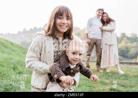 Eine weiße Teenagerin mit braunen Augen, dunklen Haaren und Pony in einem Kleid mit Leopardenmuster und einem Strickpullover hält ihren kleinen Bruder Kleinkind angezogen Stockfoto