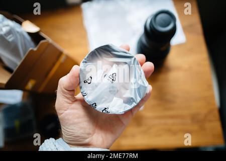 POV männliche Hand hält neue Streulichtblende im Kunststoffgehäuse während des Auspackens eines neuen Hochleistungs-Kameraobjektivs mit Kunststoffschutz Stockfoto