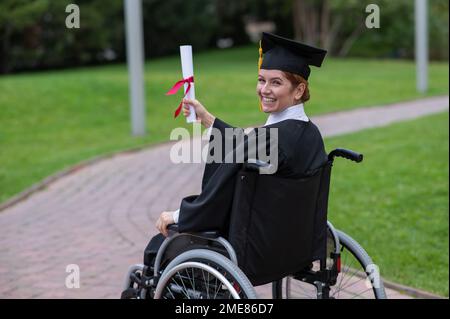 Glückliche weiße Frau im Rollstuhl dreht sich um und hält ihr Diplom draußen. Stockfoto