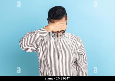 Junger erwachsener Geschäftsmann, der die Augen mit der Hand schließt, will das nicht sehen, Probleme ignorieren, sich vor stressigen Situationen verstecken, gestreiftes Hemd tragen. Innenstudio, isoliert auf blauem Hintergrund. Stockfoto