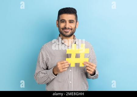 Virales Hashtag und erfolgreiches Blogging. Zufriedener Geschäftsmann mit Bart, der ein großes gelbes Hasensymbol in der Hand hält und ein gestreiftes Hemd trägt. Innenstudio, isoliert auf blauem Hintergrund. Stockfoto