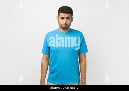 Porträt eines traurigen, verärgerten, deprimierten, unrasierten Mannes, der ein blaues T-Shirt trägt, in die Kamera schaut, mit Stirnrunzeln, Traurigkeit ausdrückt, beleidigt ist. Studioaufnahmen im Innenbereich isoliert auf grauem Hintergrund. Stockfoto