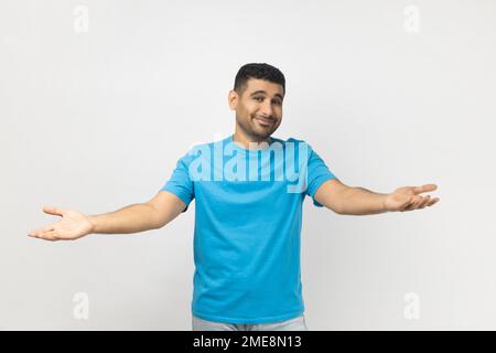 Das Porträt eines unrasierten Mannes mit blauem T-Shirt zeigt eine willkommene Geste, verbreitet Hände, wie er seinen besten Freund kuscheln will, lächelt breit. Studioaufnahmen im Innenbereich isoliert auf grauem Hintergrund. Stockfoto