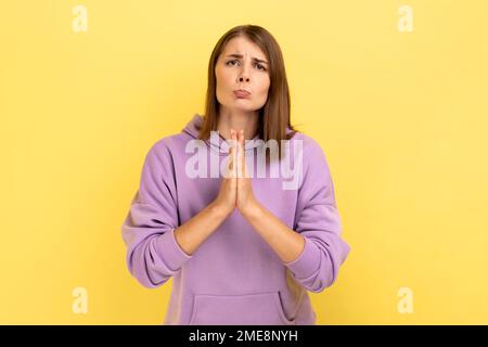 Porträt einer flehenden Frau, die die Arme in Gebetsgesten hält und um Vergebung bittet, die Fehler bemitleidet, lila Kapuzenpullover trägt. Innenstudio-Aufnahme isoliert auf gelbem Hintergrund. Stockfoto