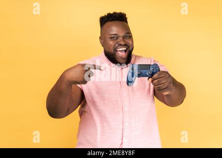Porträt eines fröhlichen, positiven, bärtigen Mannes, der ein rosa Hemd trägt, mit aufgeregtem Ausdruck steht und auf den Joystick zeigt. Innenstudio-Aufnahme isoliert auf gelbem Hintergrund. Stockfoto