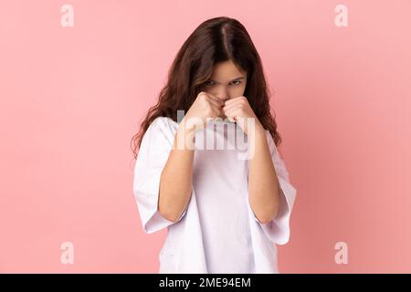 Porträt eines besorgten kleinen Mädchens, das ein weißes T-Shirt trägt und geballte Fäuste zum Boxen hält, Selbstverteidigungskurse für Kinder, Kampfgeist. Studio-Aufnahme im Innenbereich isoliert auf pinkfarbenem Hintergrund. Stockfoto