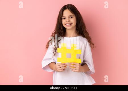 Porträt eines niedlichen kleinen Mädchens, das ein weißes T-Shirt trägt, das Hashtag-Symbol in der Hand hält und mit einem zahnigen Lächeln in die Kamera schaut und ein Hash-Schild zeigt. Studio-Aufnahme im Innenbereich isoliert auf pinkfarbenem Hintergrund. Stockfoto