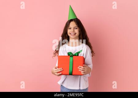 Porträt eines kleinen Mädchens mit weißem T-Shirt und Partykegel in der rot verpackten Geschenkbox, das mit Zufriedenheit in die Kamera schaut. Studio-Aufnahme im Innenbereich isoliert auf pinkfarbenem Hintergrund. Stockfoto