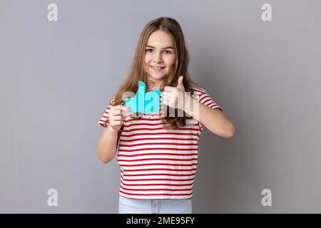 Ein kleines Mädchen trägt ein gestreiftes T-Shirt, das die Daumen hoch hält, ein blaues Symbol, ein Symbol für Follower-Benachrichtigungen, Empfehlungen und gutes Feedback in sozialen Medien. Studioaufnahmen im Innenbereich isoliert auf grauem Hintergrund. Stockfoto