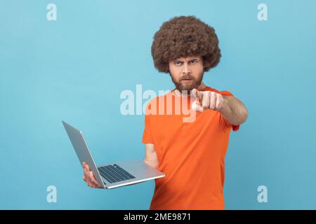Strenger, herrisch gestalteter Mann mit Afro-Haarschnitt, der ein orangefarbenes T-Shirt trägt, den Laptop in den Händen hält, online arbeitet, mit dem Finger auf die Kamera zeigt, mit ernstem Gesichtsausdruck. Innenstudio, isoliert auf blauem Hintergrund. Stockfoto